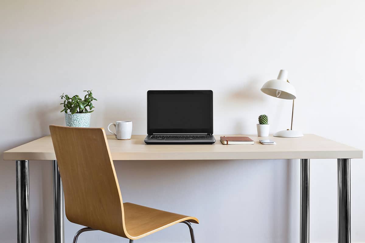 A minimalist study desk setup with a laptop, wooden chair, and desk lamp against a clean white wall.