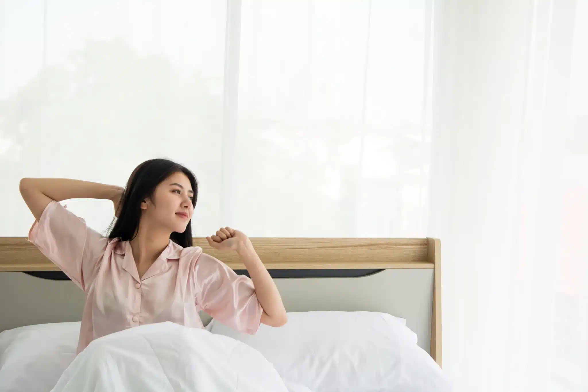 A woman stretching while waking up on a sturdy wooden modern bedframe malaysia in a bright bedroom.