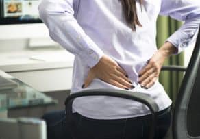 An office worker in pain clutches their lower back while sitting at a desk, highlighting the need for a supportive ergonomic chair in Malaysia.