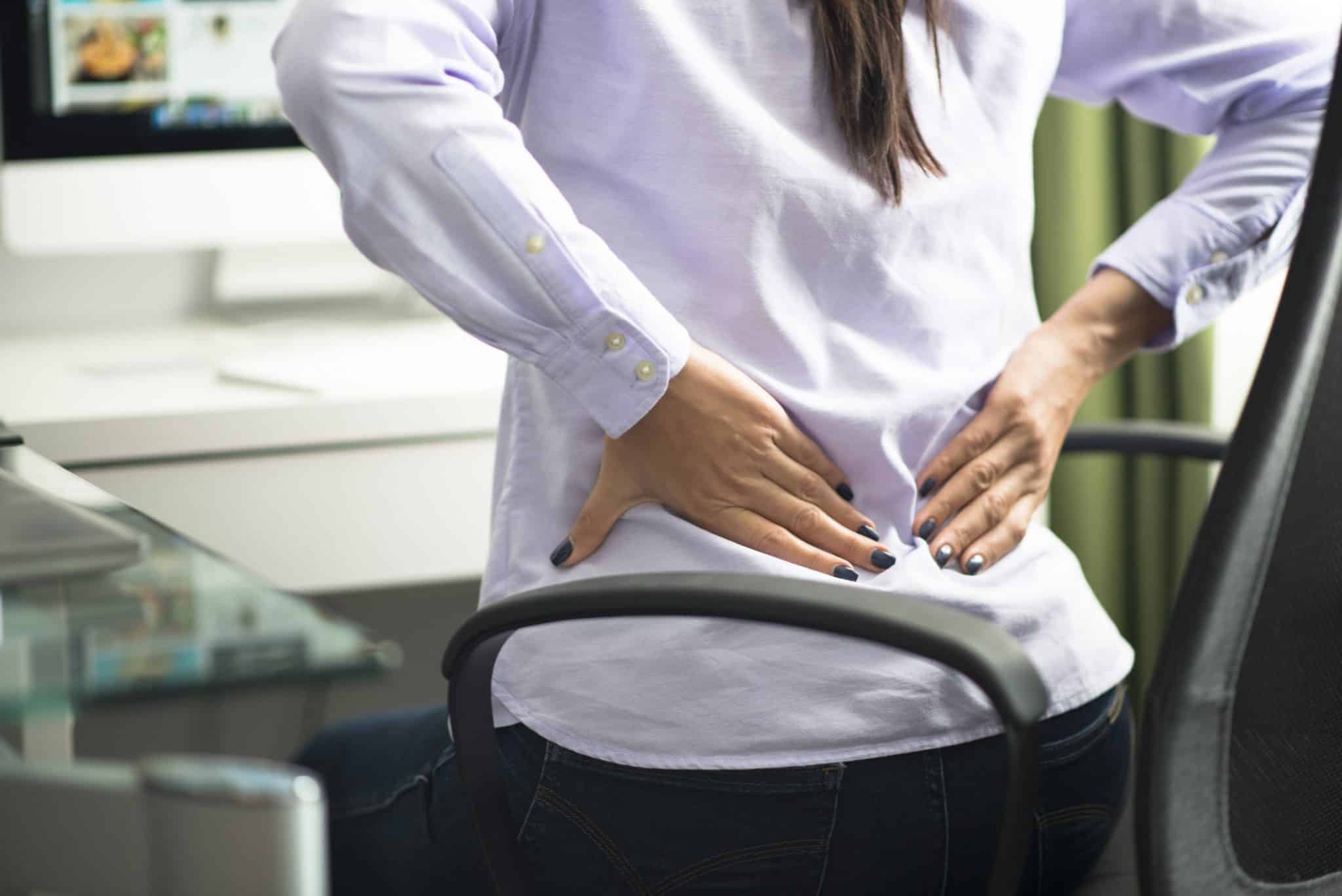 An office worker in pain clutches their lower back while sitting at a desk, highlighting the need for a supportive ergonomic chair in Malaysia.